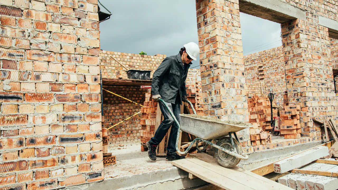 Builder pushing a wheelbarrow along a plank inside the brick shell of a new house