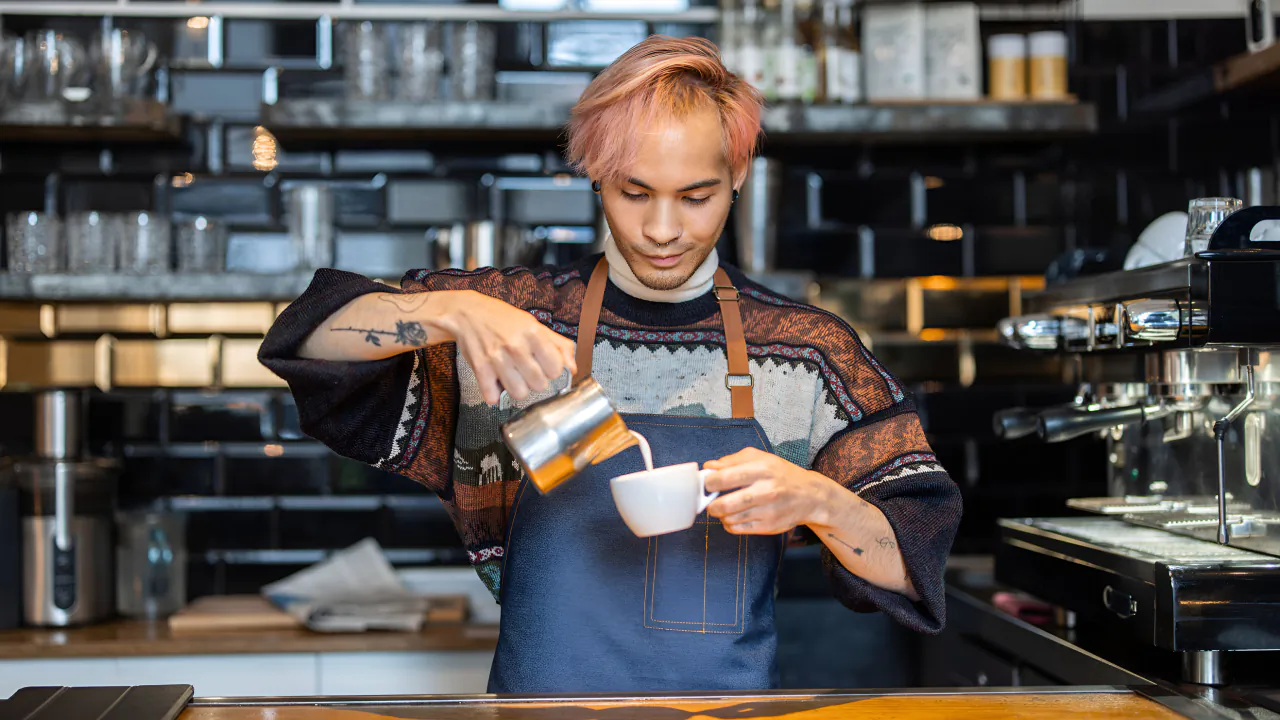 Barista in a denim apron pouring steamed milk into a cup behind an independent coffee shop counter