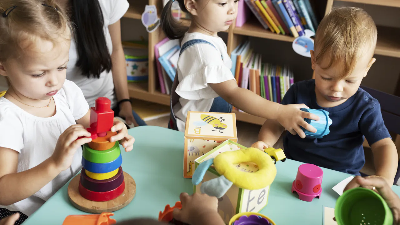 Nursery children building with colourful toys at a low table while a practitioner helps