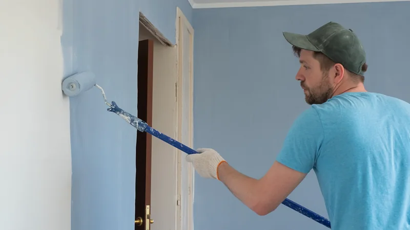 Decorator rolling pale blue emulsion onto a living-room wall with an extension pole