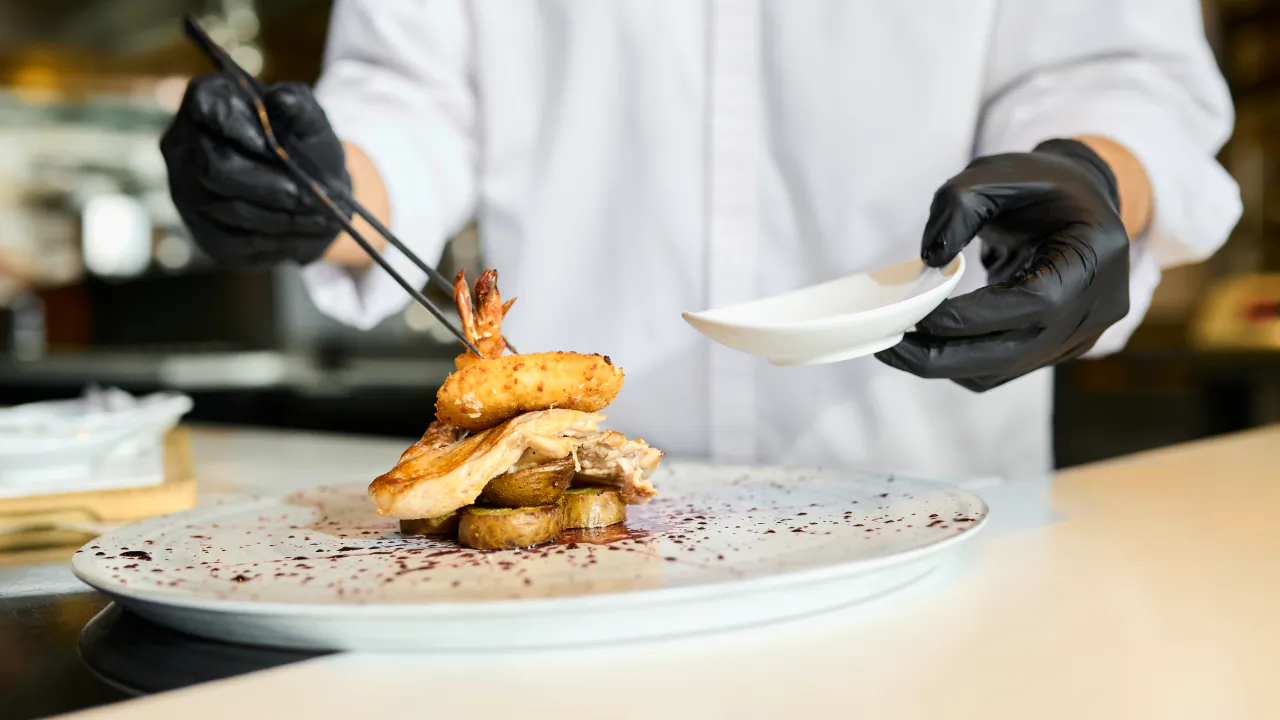 Chef in black gloves finishing a plate of food at the pass of an open restaurant kitchen