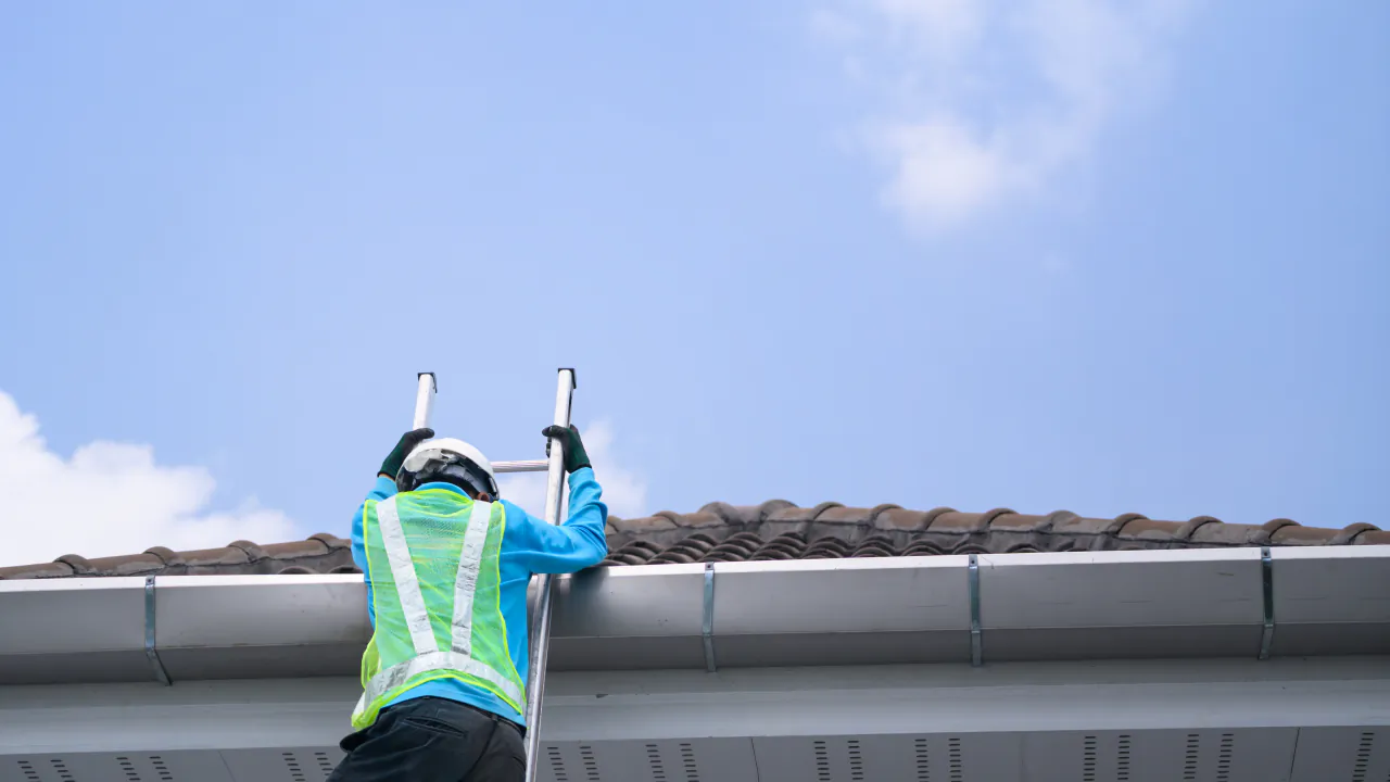 Roofer in a hi-vis vest climbing a ladder to the eaves of a tiled roof under a blue sky