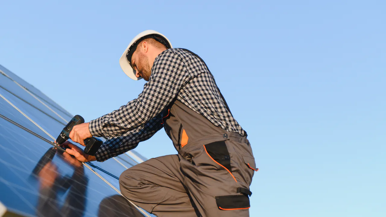 Solar installer in a hard hat fixing a photovoltaic panel to a roof against a blue sky