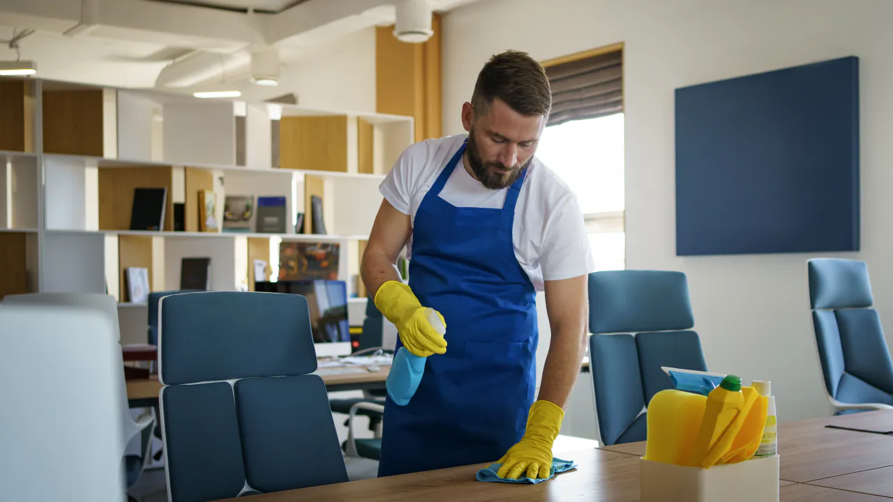 Cleaner in an apron and gloves wiping down an office desk with a spray and cloth
