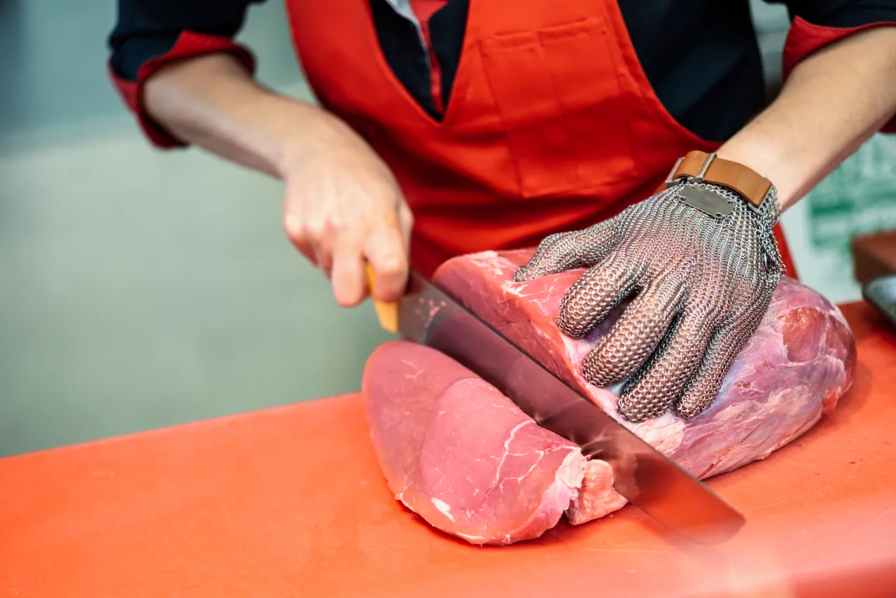 A butcher in a red apron and mesh glove cutting a large joint of fresh meat on a board