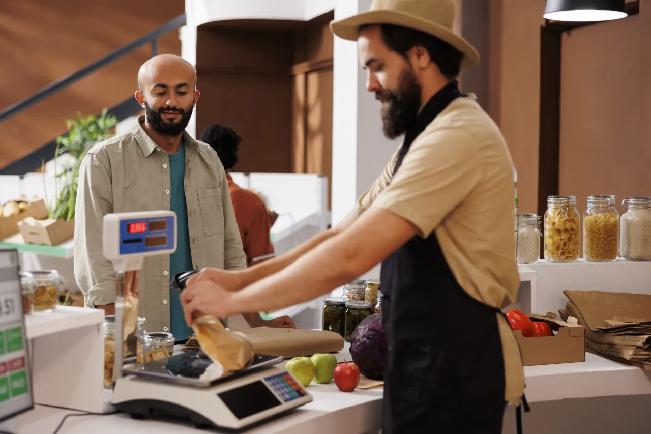 Two shopkeepers in a small food shop weighing a loaf on a scale for a customer