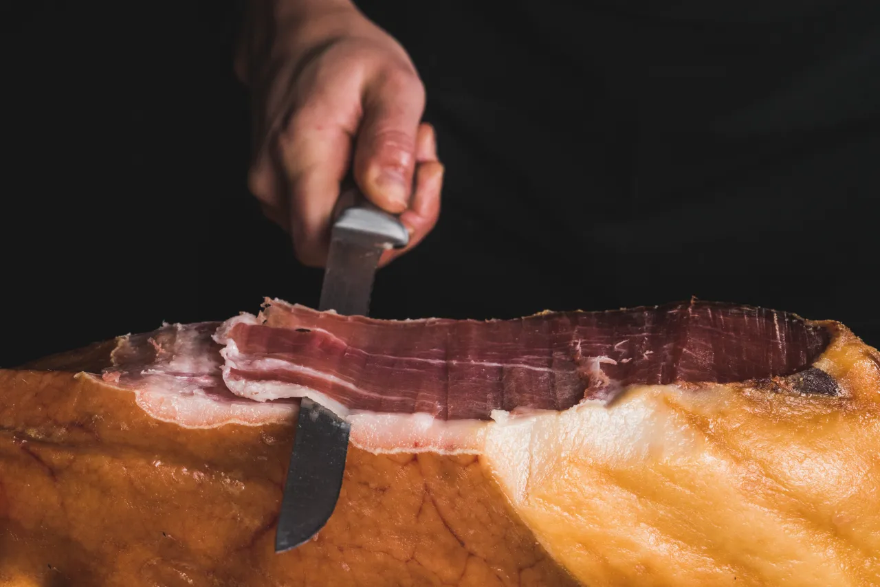 Hands carving thin slices from a cured ham leg with a long knife