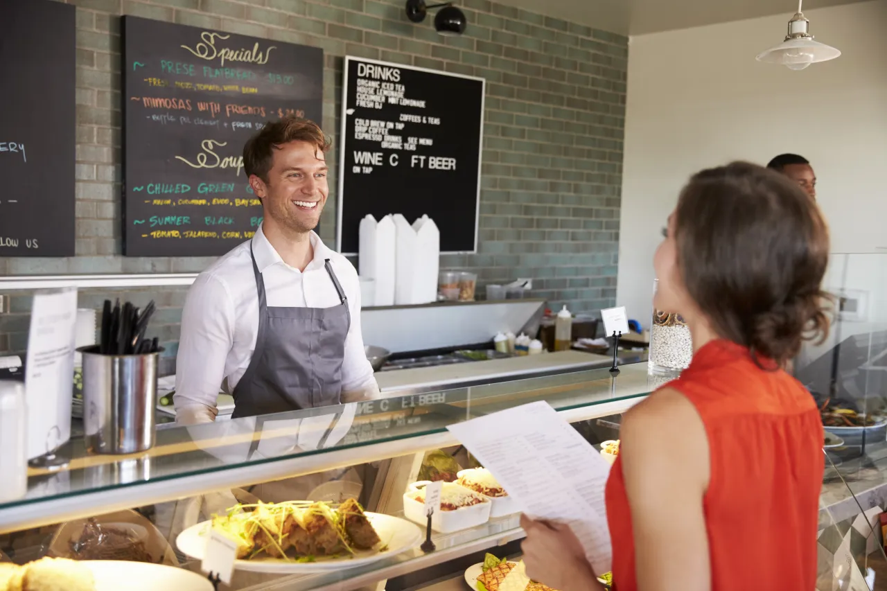 Deli owner smiling behind a counter beneath a chalkboard menu, serving a customer