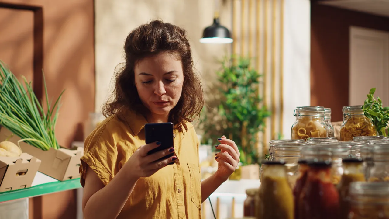 Woman checking her phone beside shelves of jars and fresh produce in an independent food shop
