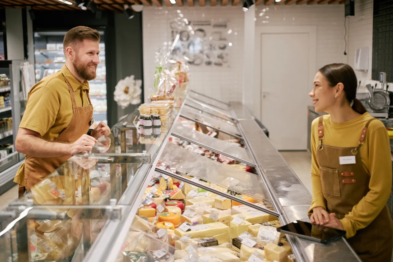 Two deli staff in aprons chatting across a glass counter of cheeses and cooked foods