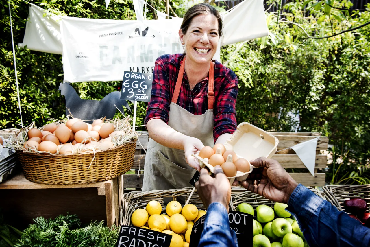 Smiling stallholder in an apron handing a box of eggs to a customer beside baskets of onions and fruit