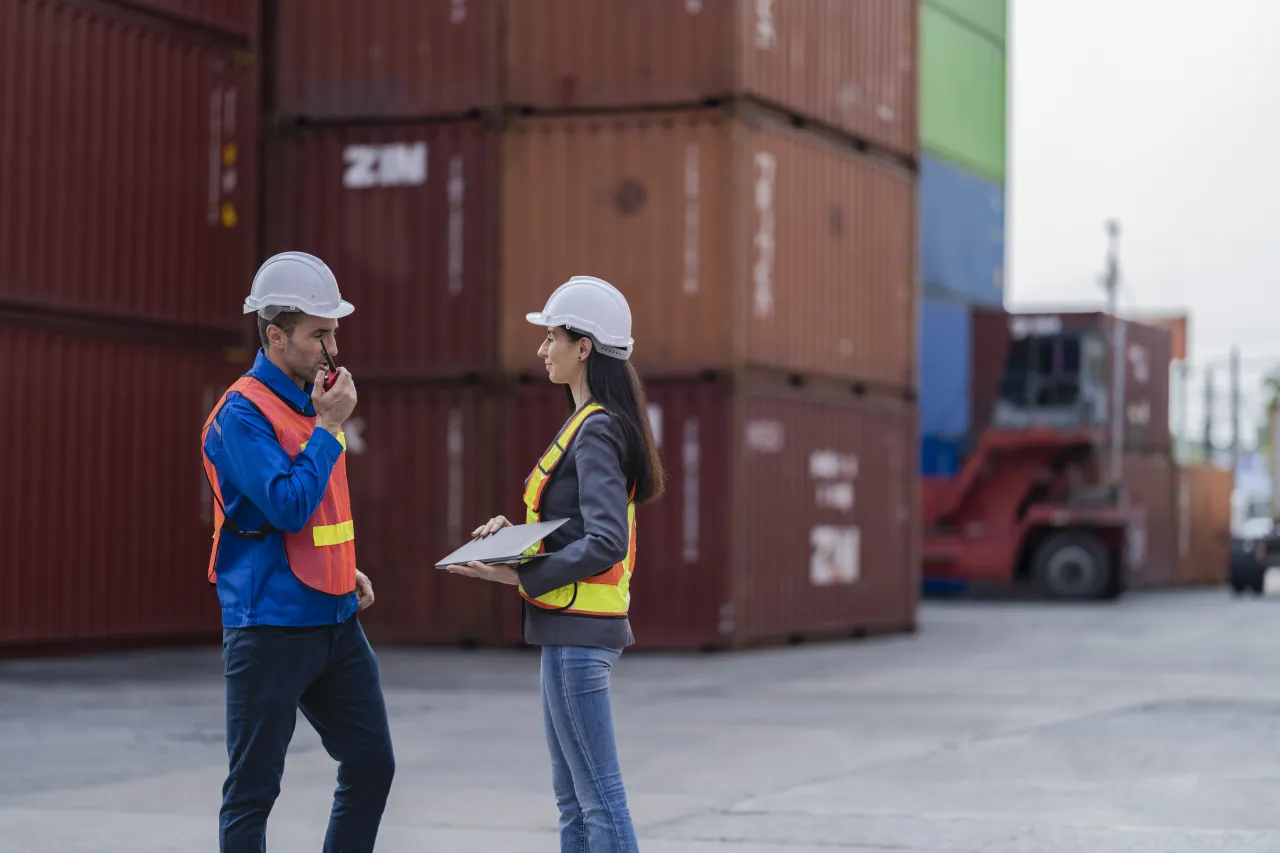 Two freight forwarding staff in hi-vis discussing a shipment's questions at a container terminal