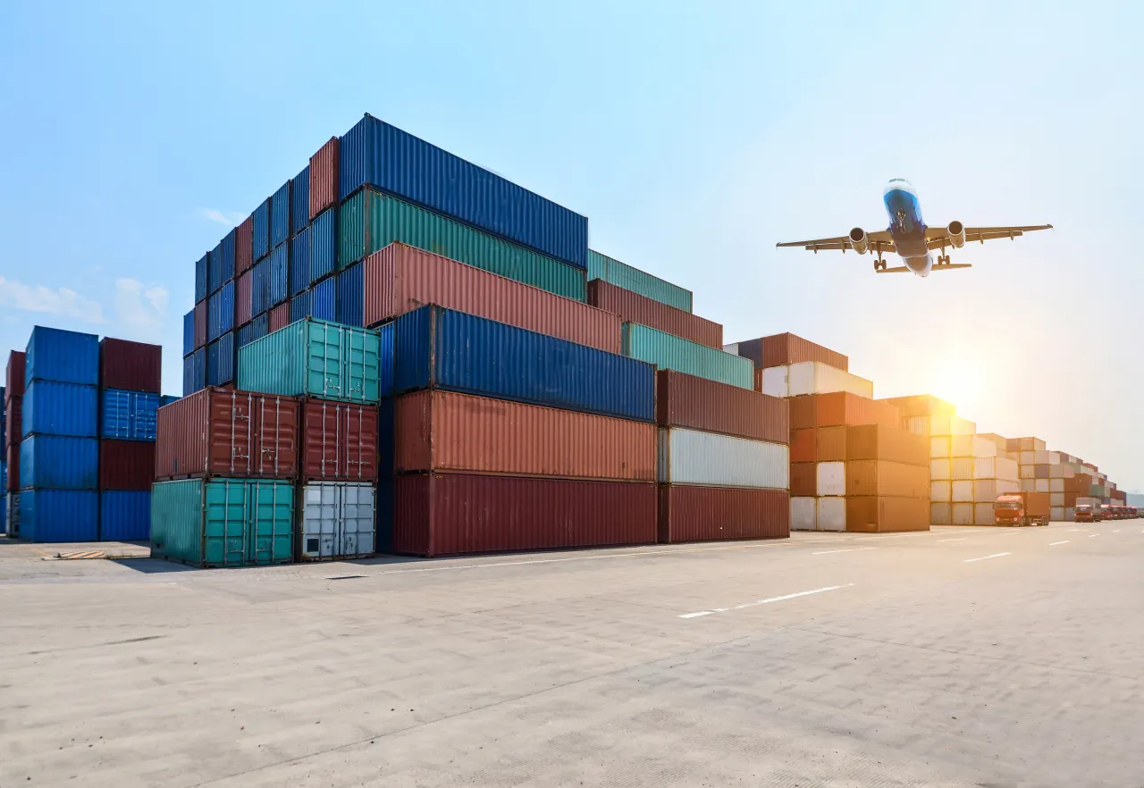 Stacked shipping containers at a freight terminal with a cargo plane overhead, representing sea and air freight forwarding