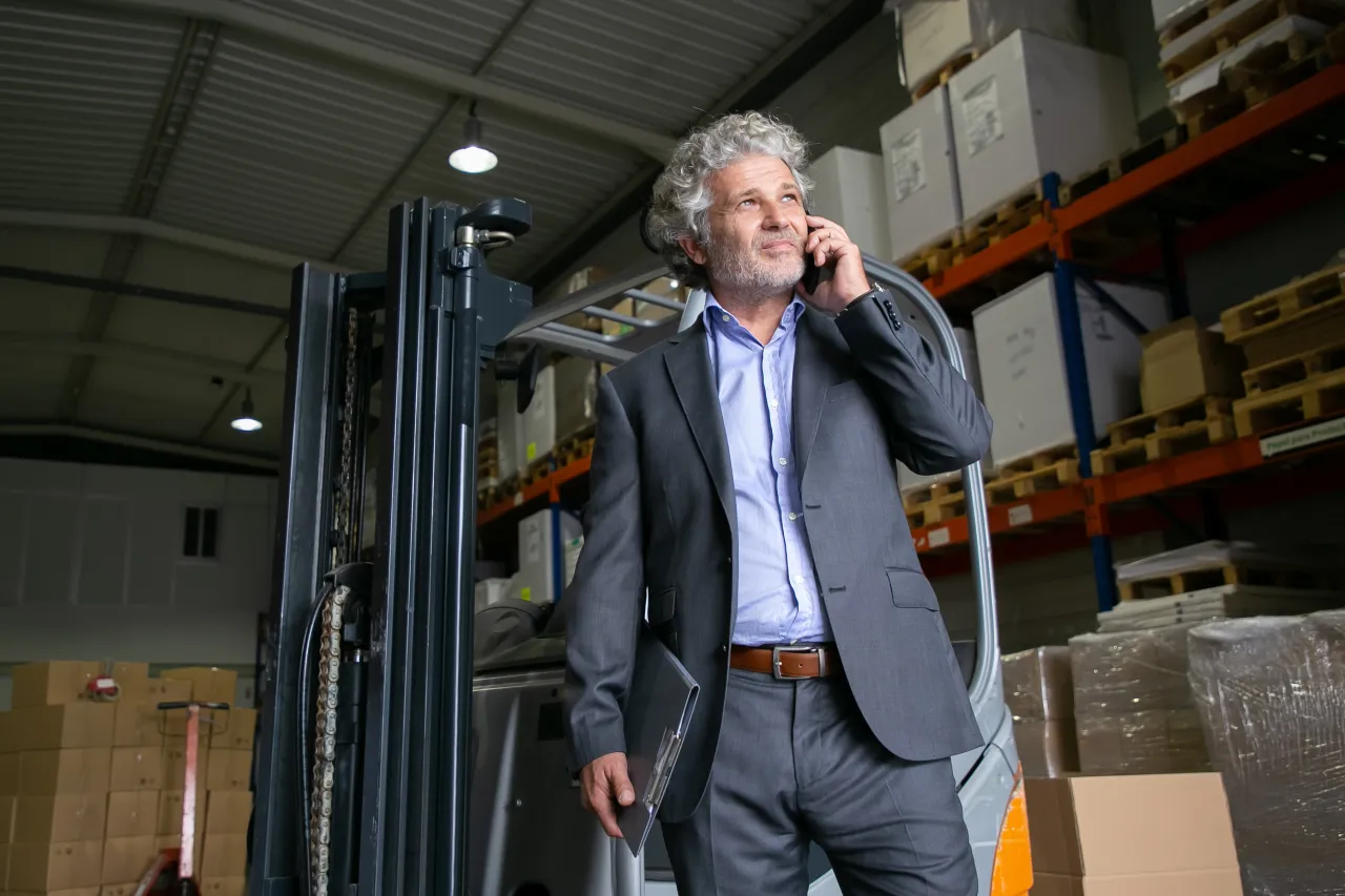 Manager in a haulage warehouse taking a call about a load to place, forklift and pallet racking behind him