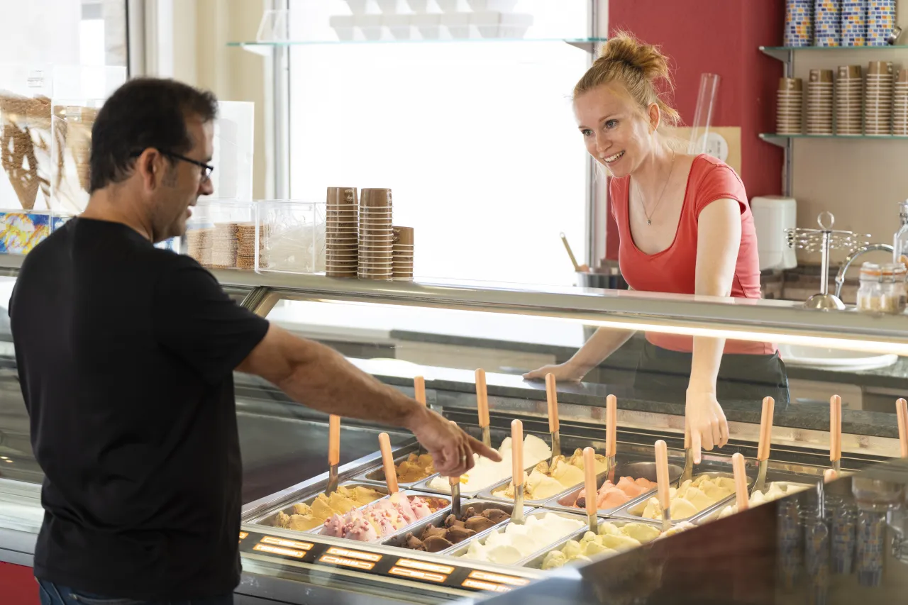 Two staff at a gelato display counter serving a customer, with stacks of cones behind them