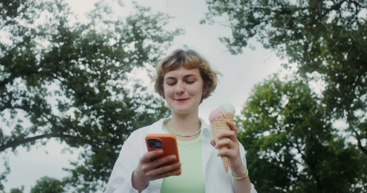 Young woman holding an ice cream cone while checking her phone under trees