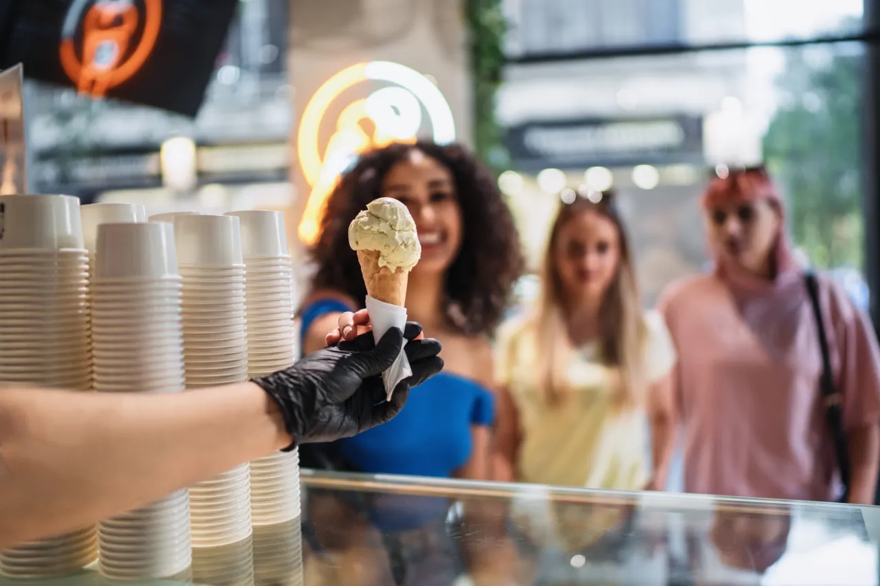 Ice cream seller in a black glove handing a cone to a group of smiling customers at the counter