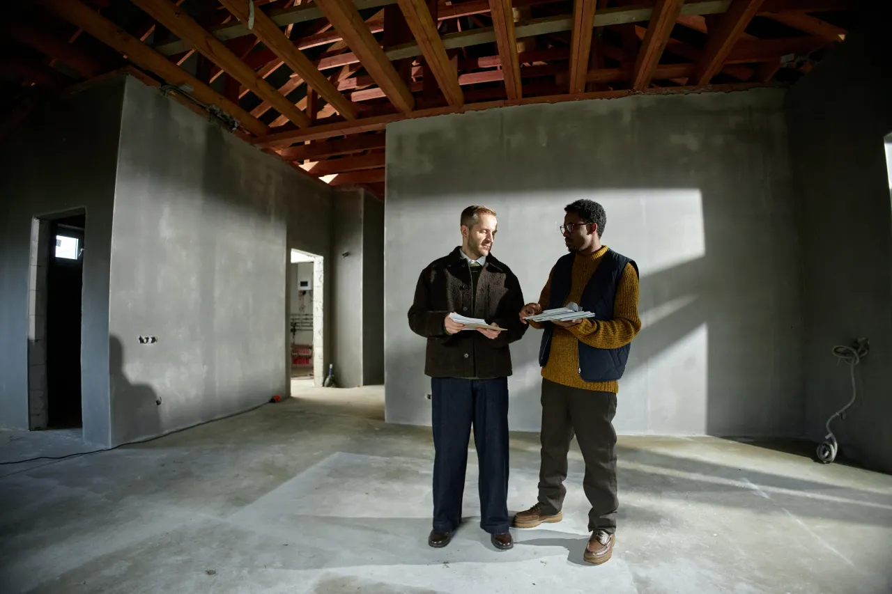 A homeowner and a professional discuss structural plans inside a house extension mid-build, exposed block wall and timber roof frame visible