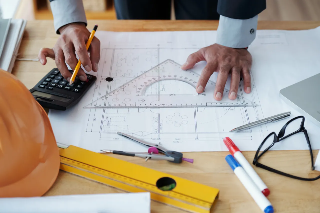 Close-up of a structural engineer using a calculator and set square while marking up a technical drawing, hard hat on the desk
