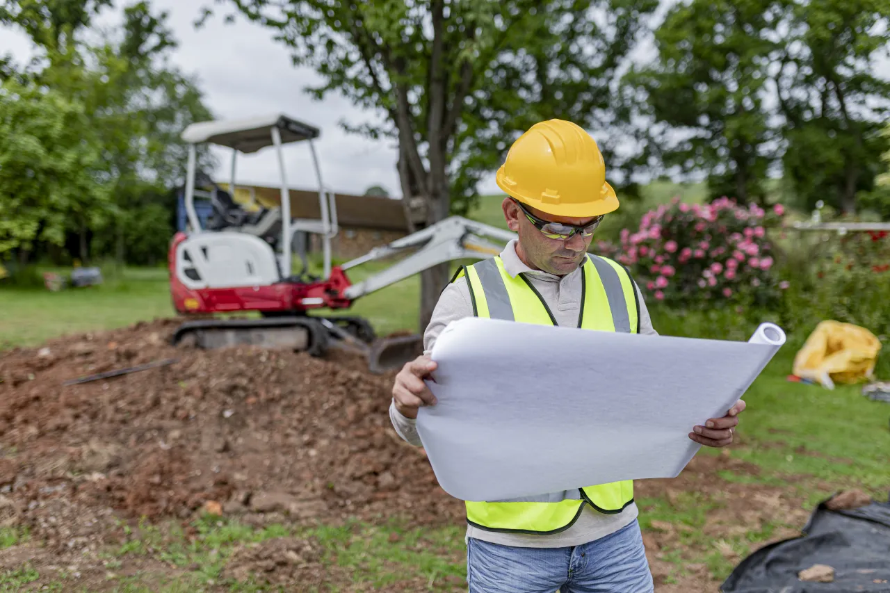 A site worker in high-vis reviews the building plan beside an excavator on a garden dig, the regulated groundwork stage of a pool build