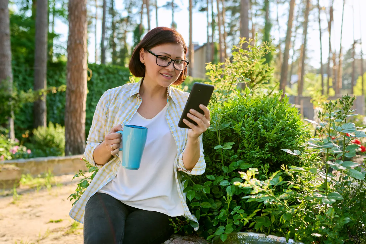 A woman sitting in her garden with a coffee, looking something up on her phone as she plans a project