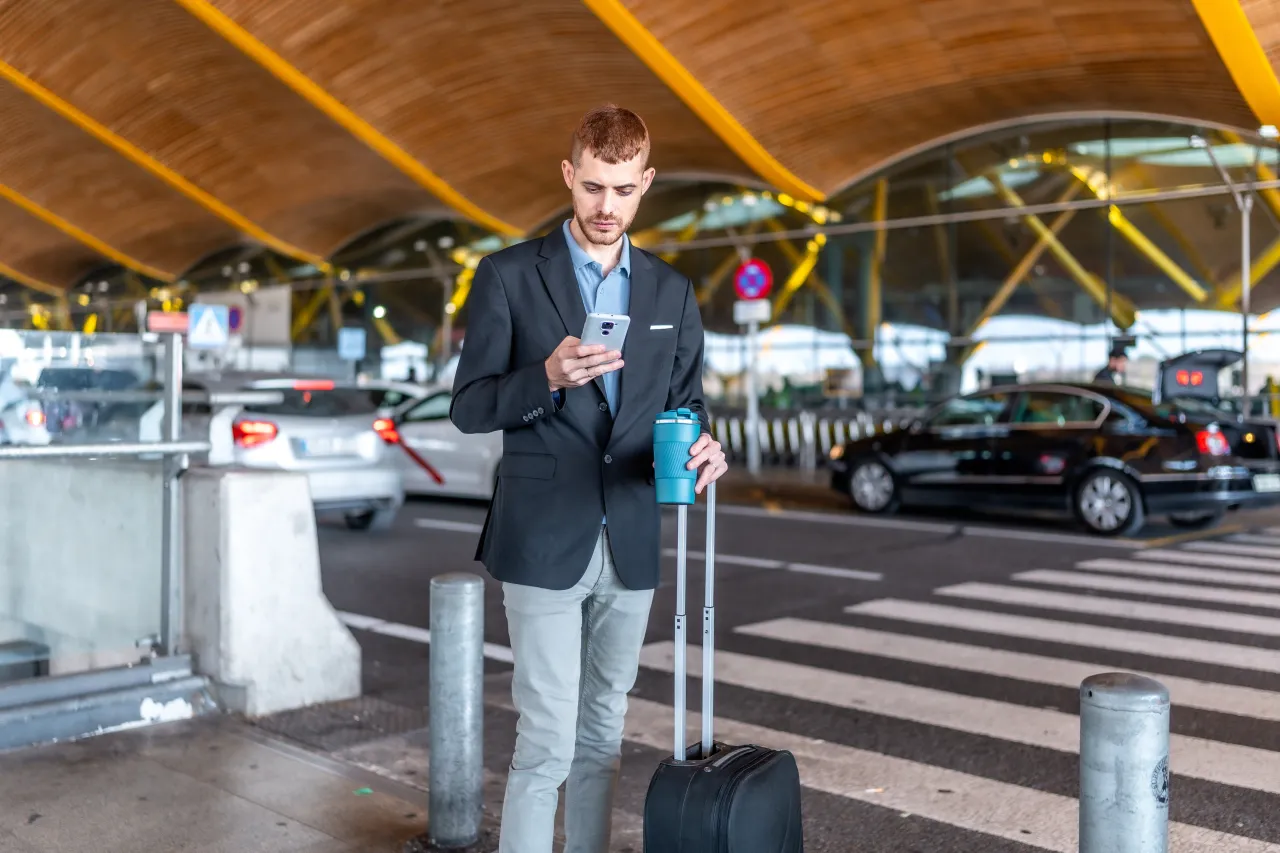 A traveller with a suitcase checking his phone at an airport kerb, cars waiting alongside
