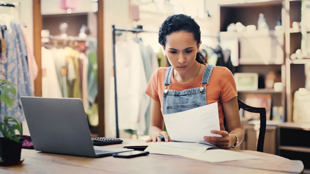 A shop owner reading paperwork at a laptop in her clothing store