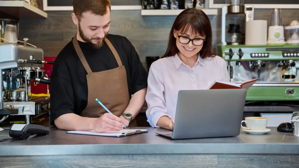Two people talking through notes and a laptop at a coffee shop counter