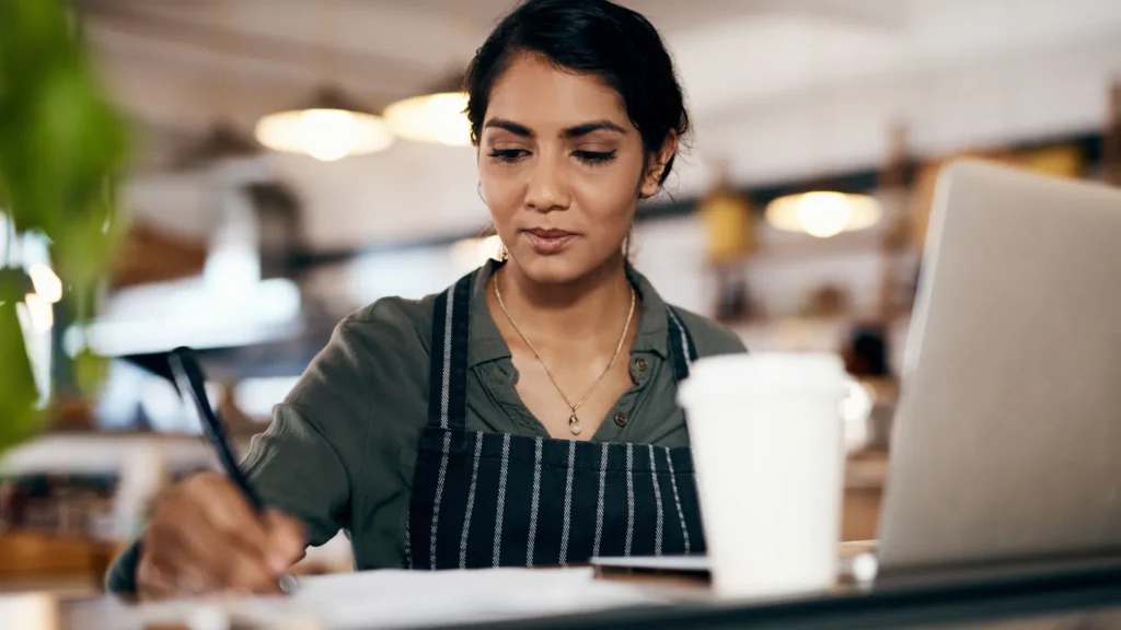 A cafe owner working through costs with a notebook and laptop at the counter