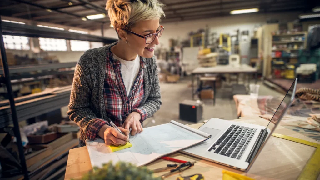 A workshop owner reviewing plans beside a laptop at her bench